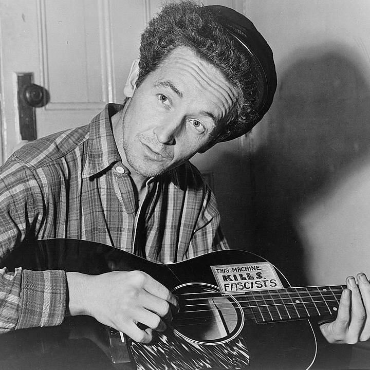 A black and white photo of Woodie Guthrie playing the guitar. On his guitar a sticker reads "This Machine Kills Fascists."