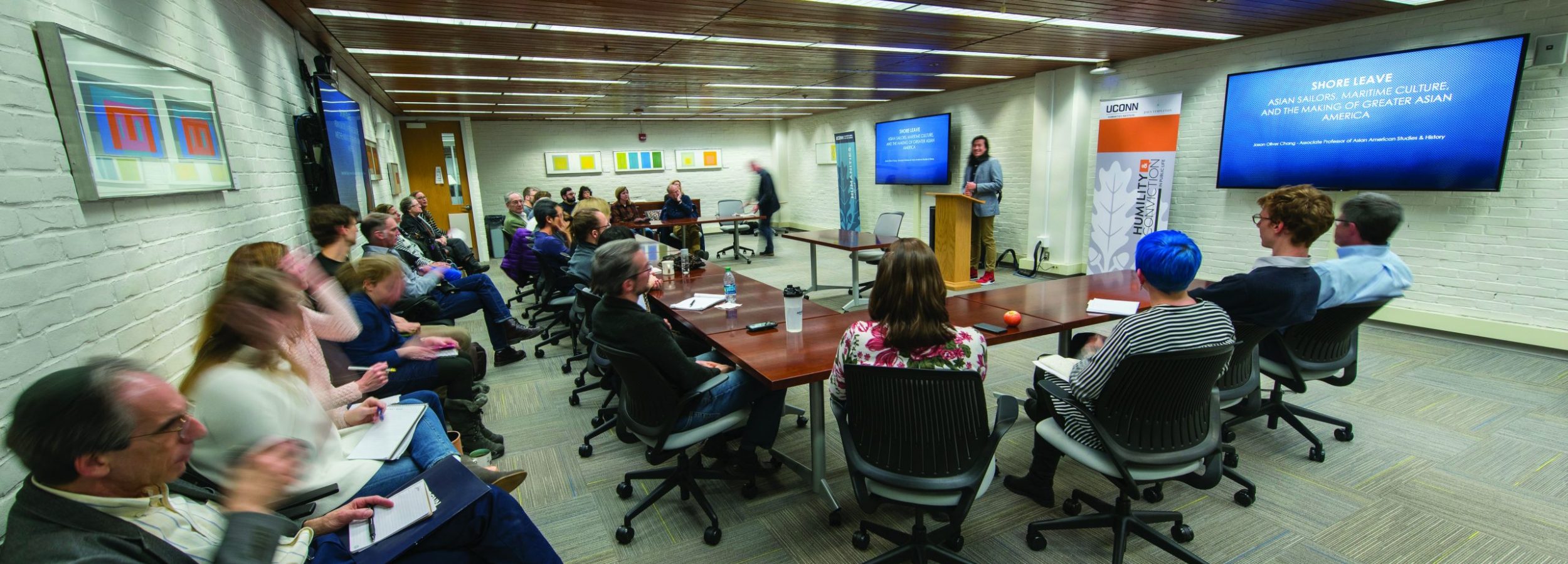A large group of faculty members and students gathered in the UCHI conference room to participate in a public event. One group sits around a conference table, while other participants sit in chairs lining the wall.