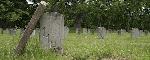 A photograph of grave stones in a green fields. The stone closest to the viewer is marked only with a series of numbers. One gravestone, broken at the base, is leaned up against its neighbour.