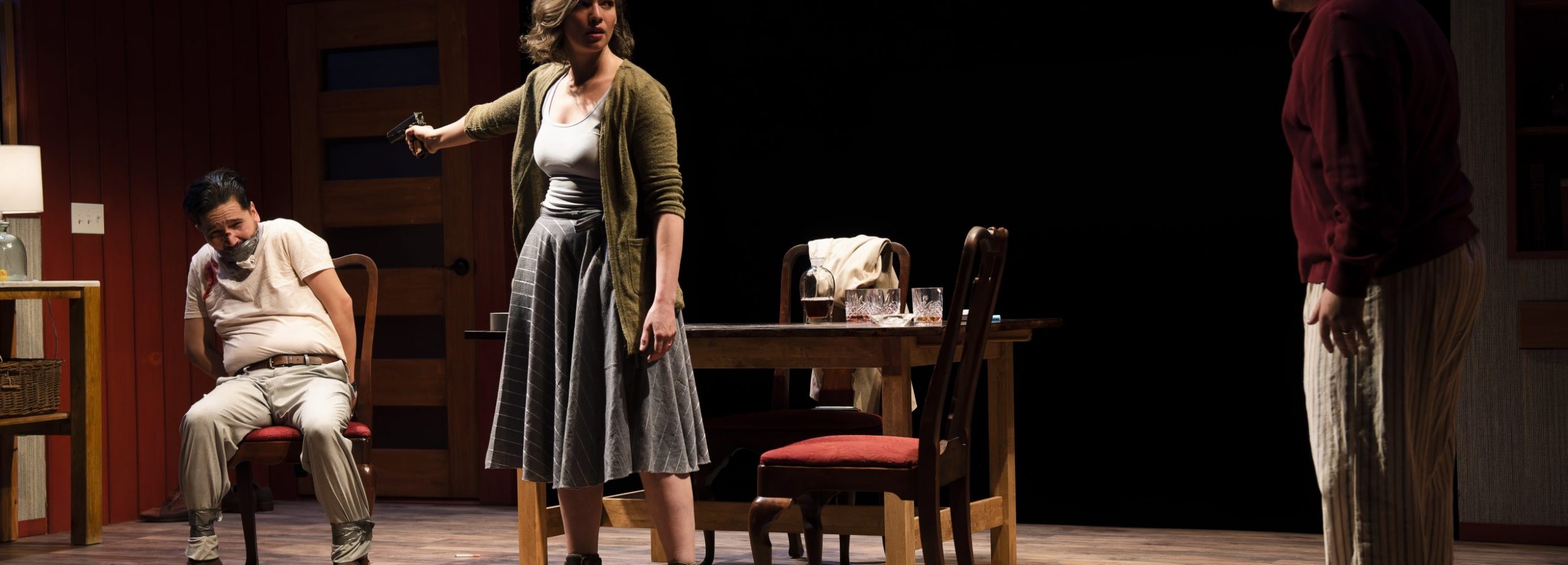 A photograph of three actors on stage, two facing one another. One of those actors is pointing a gun at the third actor, who is seated behind her, and is gagged and appears to be tied to a chair.