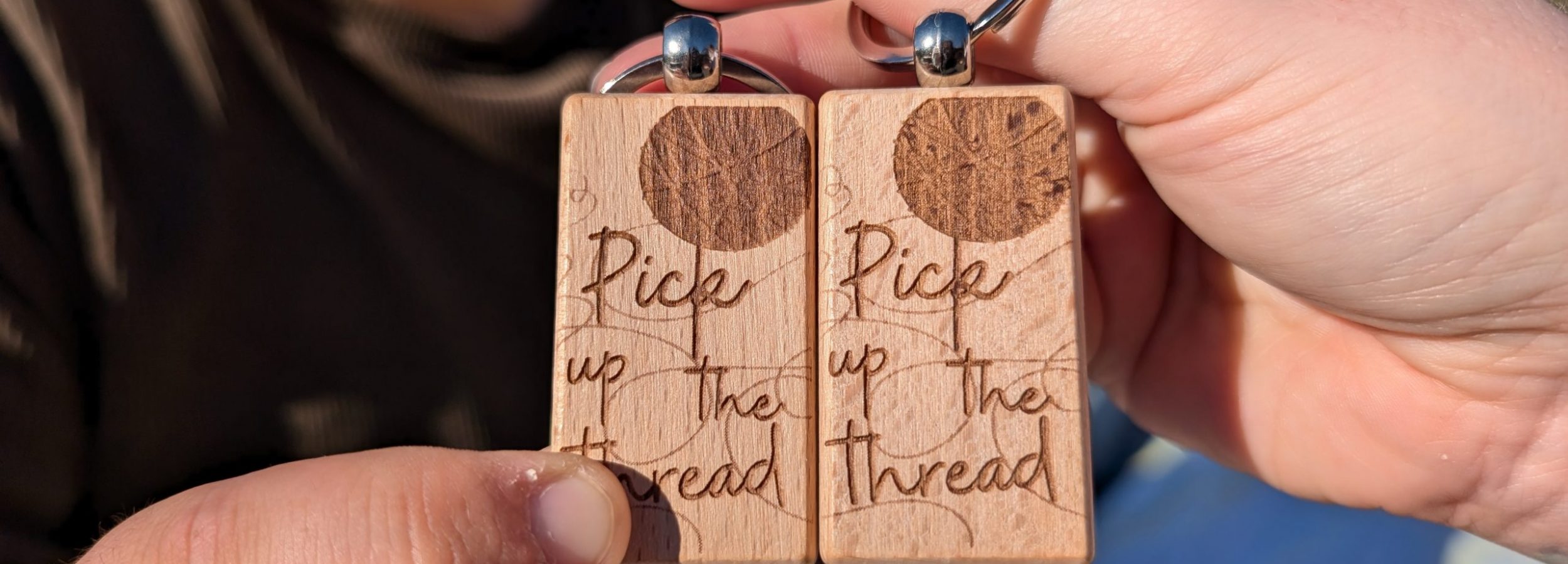 A close up photograph of two people holding wood keychains that say "Pick Up the Thread."